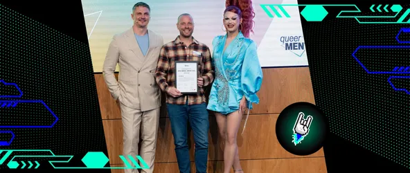 Preisträger Nils Binder hält die Urkunde des Queermentor Role Model Awards in der Hand und lächelt in die Kamera. Neben ihm stehen die Moderator*innen der Award-Show Rachel Intervention und Pavlo Stroblja.